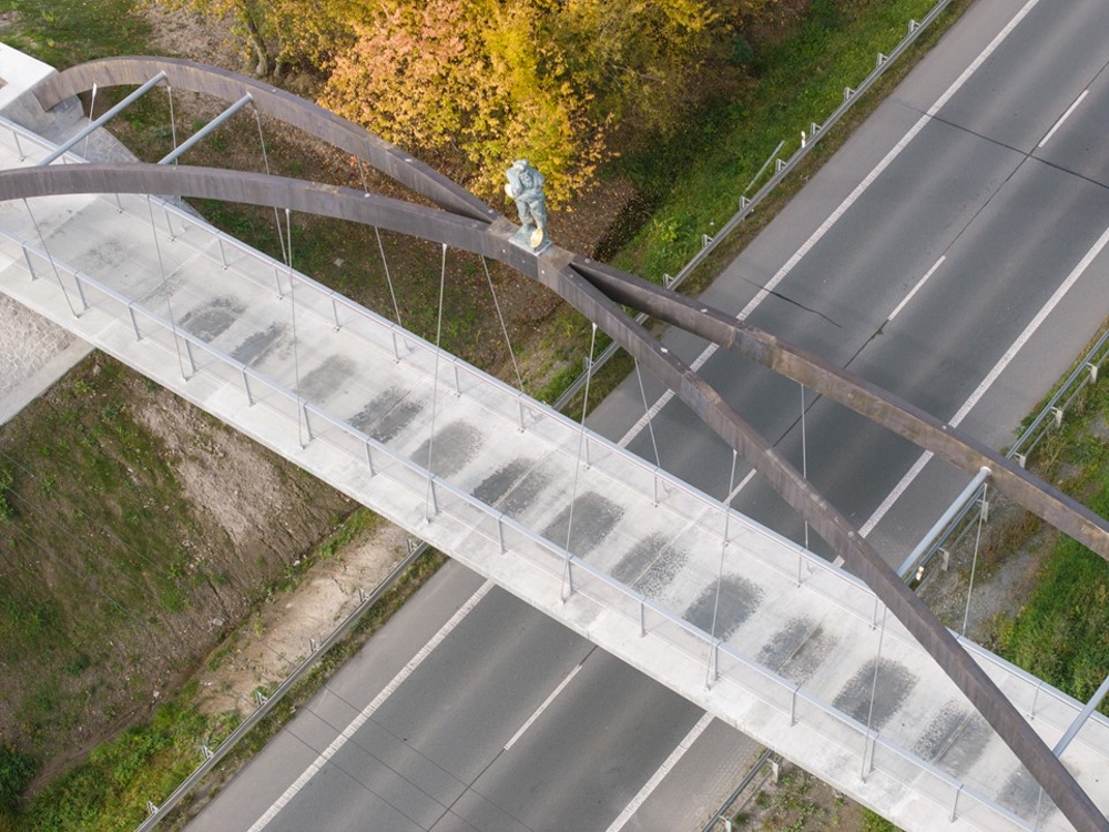 Person wearing yellow jacket walks on a curved metal pedestrian bridge above a road from an aerial view