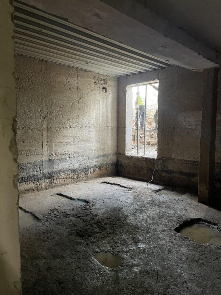 Concrete-walled room under renovation with exposed pipes on the ceiling and holes in the floor; a worker in a high-visibility vest is visible through a window opening outside.