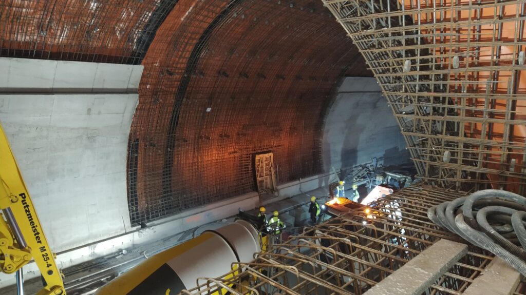 Construction workers in hard hats stand at the base of a curved tunnel wall with exposed rebar and scaffolding inside a construction site.