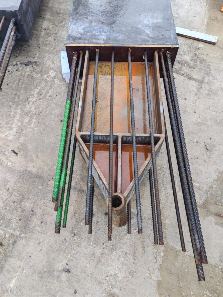 Rusty concrete formwork with vertical rebar bars around a hollow central tube on a dusty site.