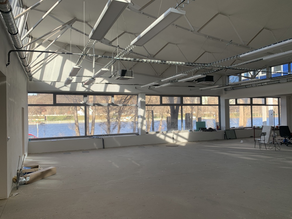 Bright, empty industrial workspace under renovation; exposed ceiling systems, large windows revealing a lake outside.
