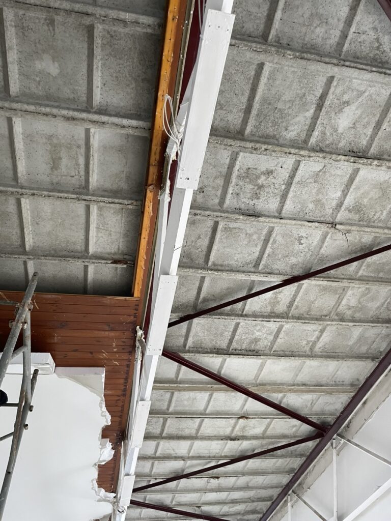 Construction site ceiling with exposed concrete slabs, red steel trusses, and a white wooden beam under renovation; scaffolding and peeling plaster visible on wall.