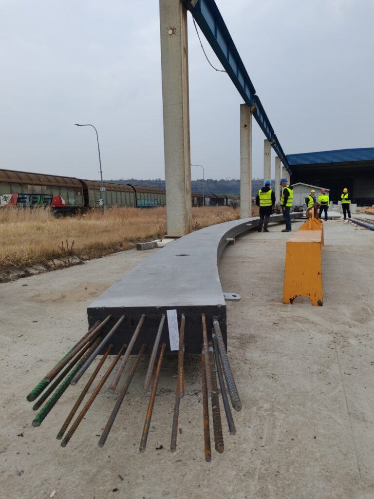 Construction crew in high-visibility vests stand beside a curved concrete beam with exposed rebar at a rail construction site.