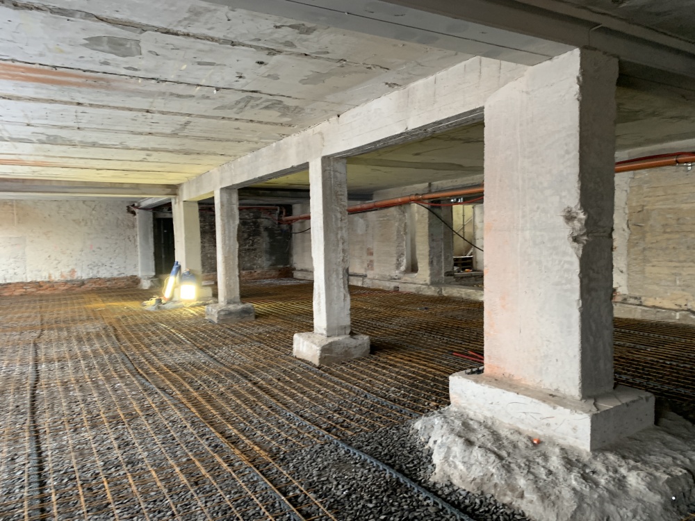 Underground construction site with concrete columns, a steel rebar grid, and two workers with bright work lights at left.