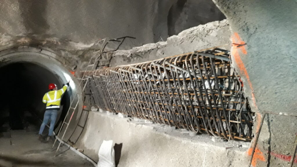 Construction worker in a yellow safety vest and red helmet standing by a large circular tunnel with exposed rebar and debris in a tunnel under construction.