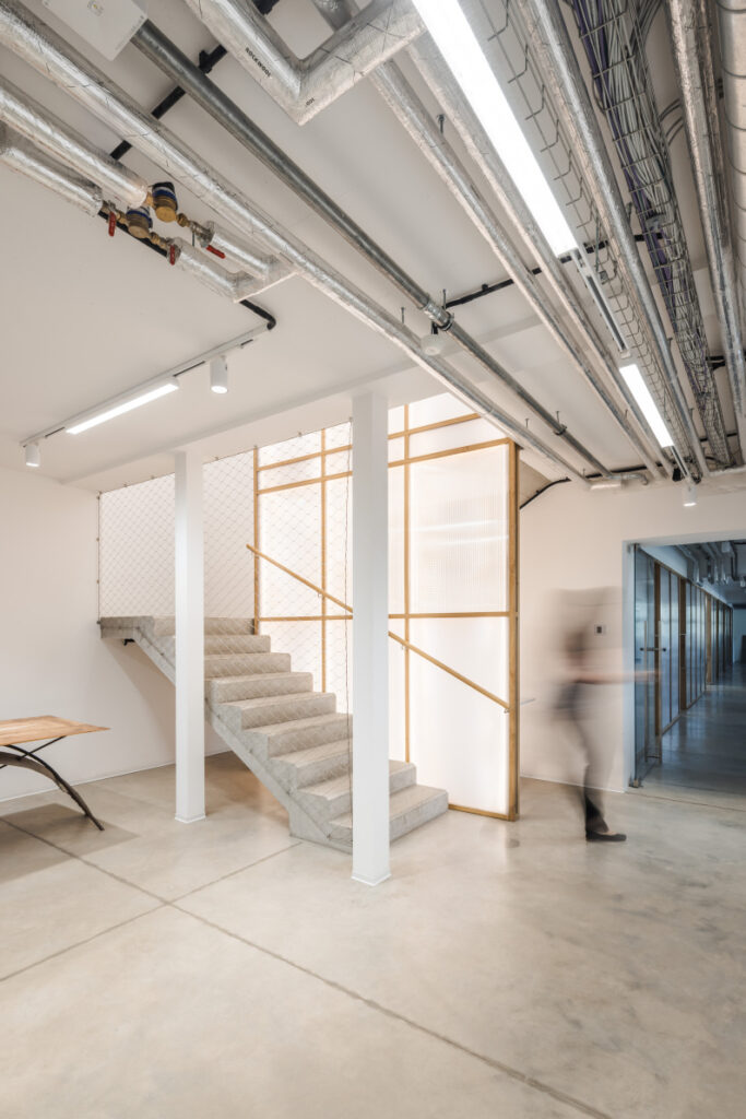Industrial interior with a concrete staircase, white columns, and exposed pipes on the ceiling; a blurred person walks along a glass-panelled corridor.