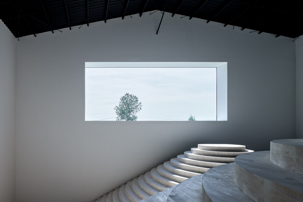 Modern interior with white walls, a rectangular window showing trees outside, and concentric stone steps formed in a circular ramp.