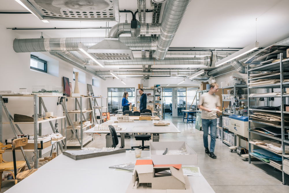 Open design studio or workshop with long tables, metal shelving, and exposed ducts; people browse materials in the background.