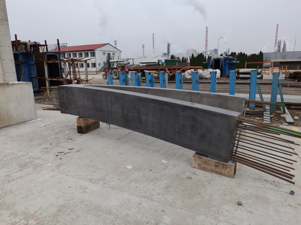 Long rectangular concrete beam resting on wooden blocks at a construction site, with exposed rebar at one end and industrial buildings in the background.