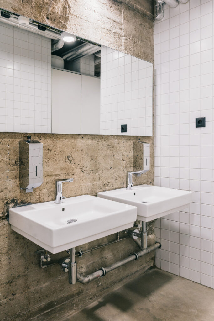 Two wall-mounted white sinks with chrome taps in a concrete-walled public bathroom, with a large mirror and soap dispenser nearby.