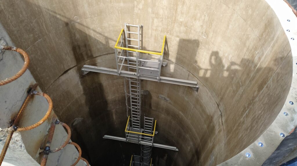 Industrial vertical shaft with metal stairway and yellow safety rails inside a circular concrete pit/tank.