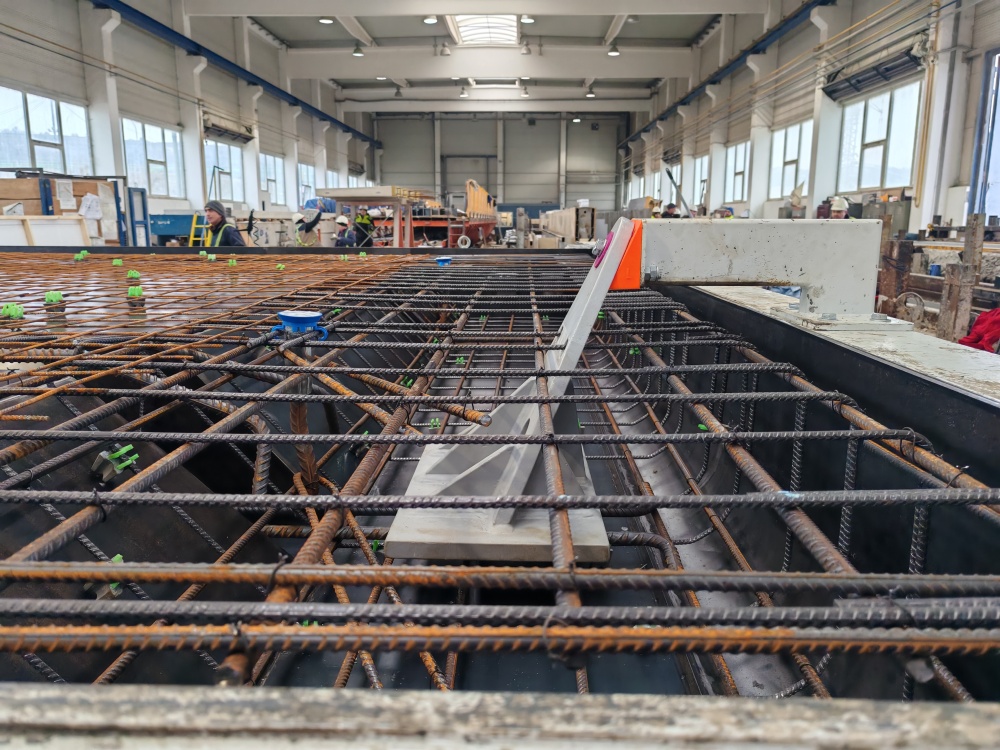 Industrial workshop with a large steel rebar grid over a pit, workers in the background near machinery.
