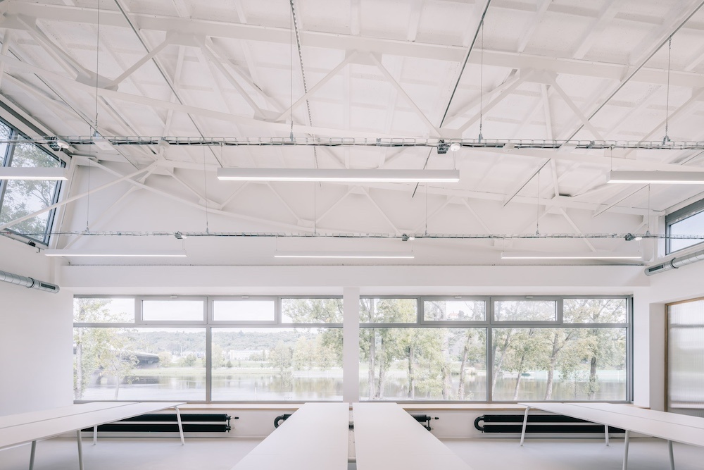 Bright, modern classroom or studio with long white tables and large windows overlooking a lake. View shows trees and water outside.
