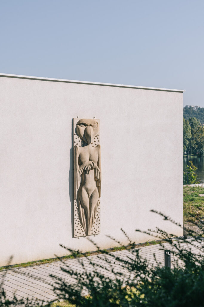 Stone relief sculpture of a stylized nude woman mounted on a pale exterior wall, with dotted border, outdoors on a sunny day.