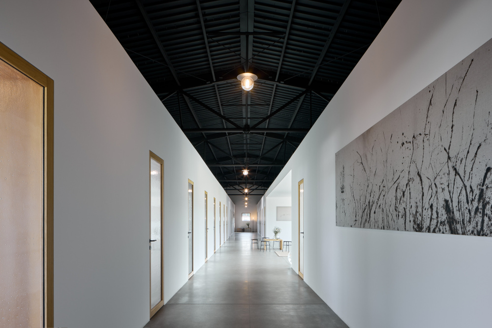 Long modern hallway with white walls, doors on both sides, and a black exposed ceiling with hanging lights; a large grayscale wall art panel runs along the right wall.