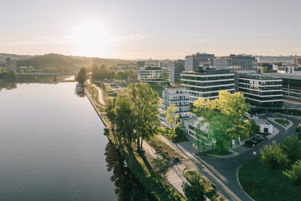 Aerial view of a riverside business district at sunset, with modern glass office towers, trees, and a curved road along the water.