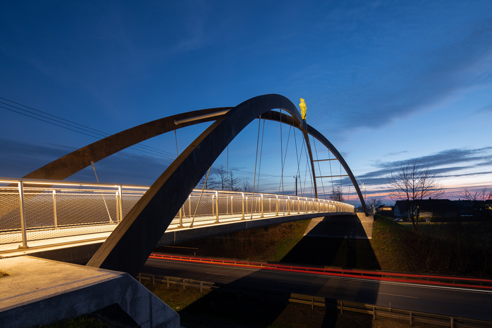 Modern arch pedestrian bridge at dusk with illuminated railing, spanning over a highway as car light trails blur below.