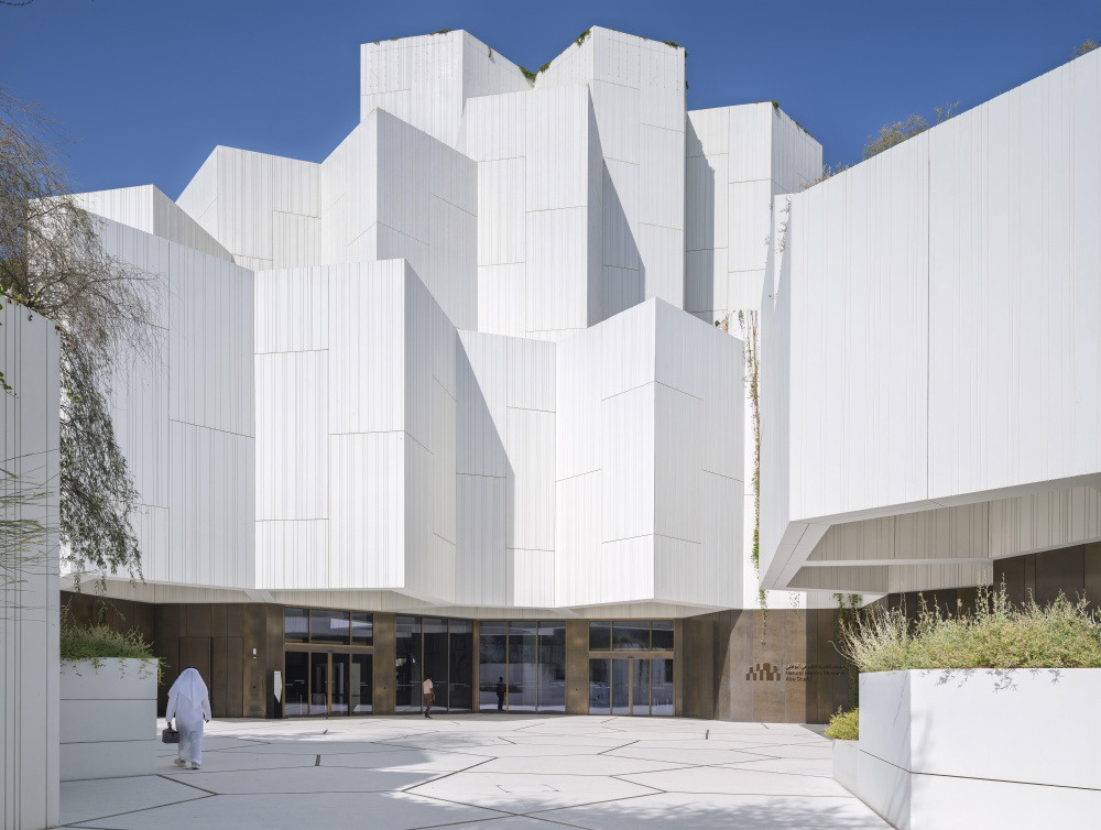 White angular modern building with blocky panels forming a sculptural facade; glass entrance doors at ground level and a person in white clothing walking across a light plaza under a blue sky.