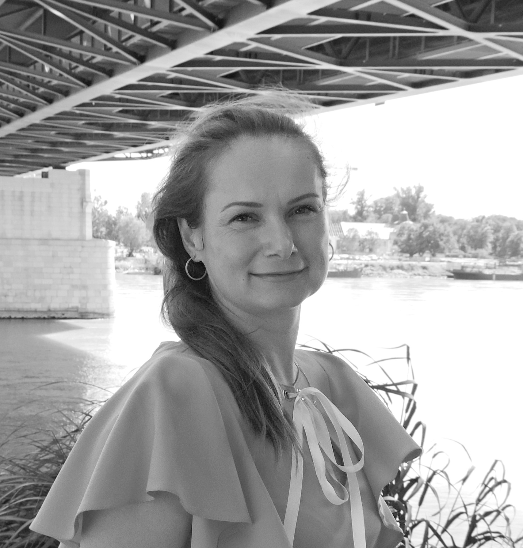 Smiling woman outdoors by a river, wearing a light blouse with a bow, under a metal bridge structure.