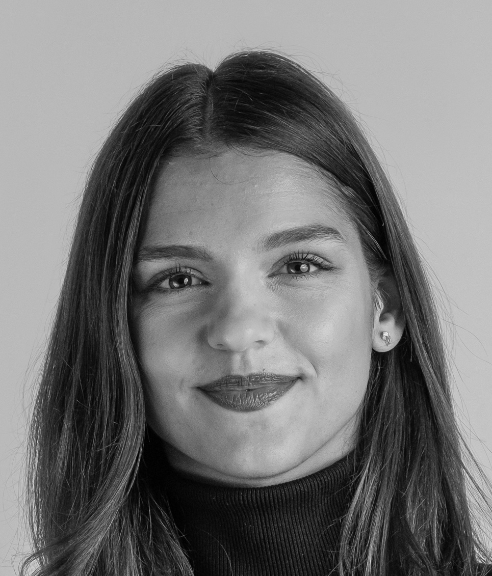 Portrait of a young woman with long dark hair smiling softly, wearing a black turtleneck against a neutral background.