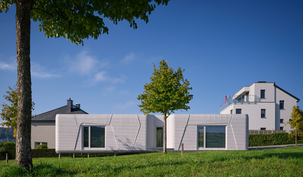 Modern white modular house on a lawn, with a single tree in front and a clear blue sky.