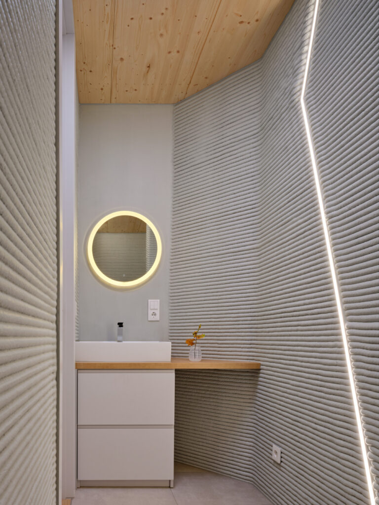 Small modern bathroom vanity with white drawers, wooden countertop, round backlit mirror, and ribbed gray walls.