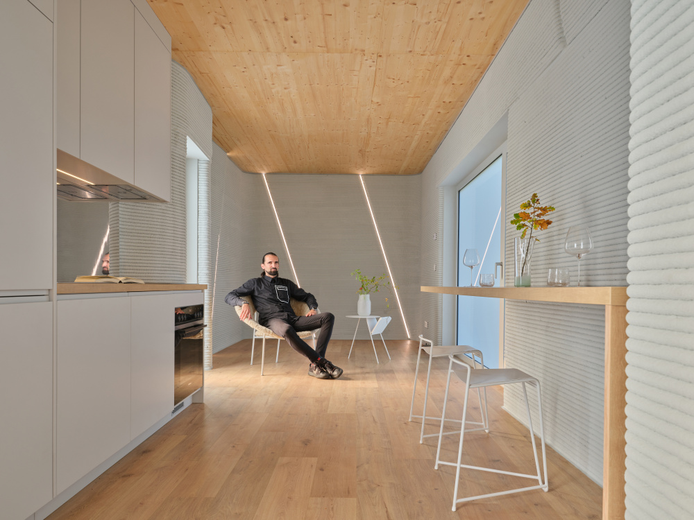 Bearded man in black sits relaxed in a minimalist white kitchen with wood flooring and a high wooden dining counter.