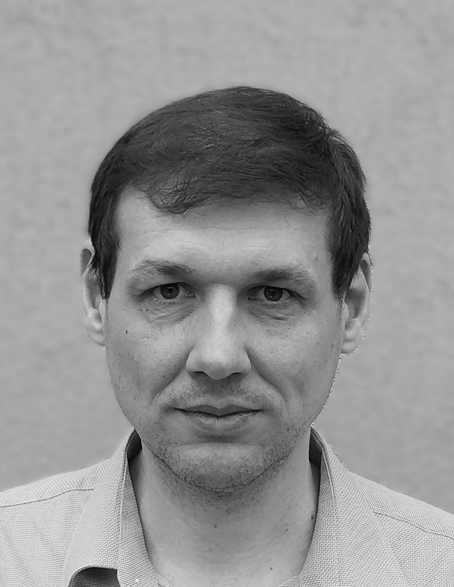 Close-up black-and-white portrait of a man with short dark hair, wearing a light checkered shirt, against a neutral backdrop.